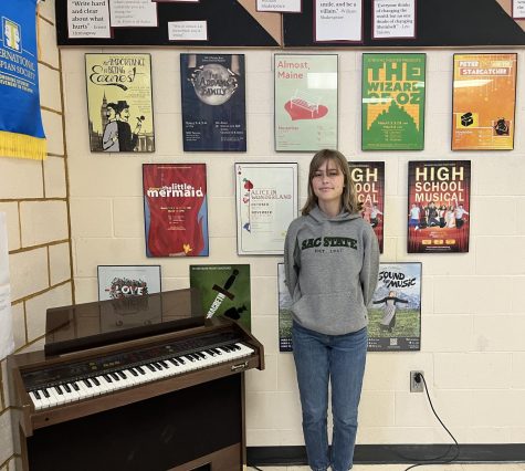 Stella Wingfield in the theater classroom in front of the past musical and show posters. Standing next to a piano, one of her favorite things.